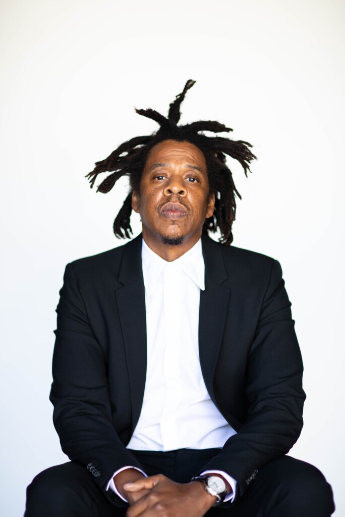 Medium shot portrait of Jay-Z. He is sitting in front of a white backdrop, wearing a black blazer, black pants, white dress shirt, and a watch. His hands are folded together in his lap.