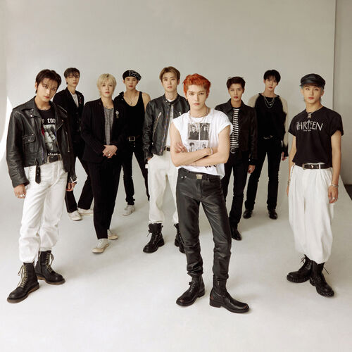 NCT 127 members standing in front of a white backdrop with natural lighting. From left: Haechan, Johnny, Yuta, Jungwoo, Jaehyun, Taeyong, Taeil, Doyoung, and Mark. They are all wearing monochromatic black-and-white outfits.