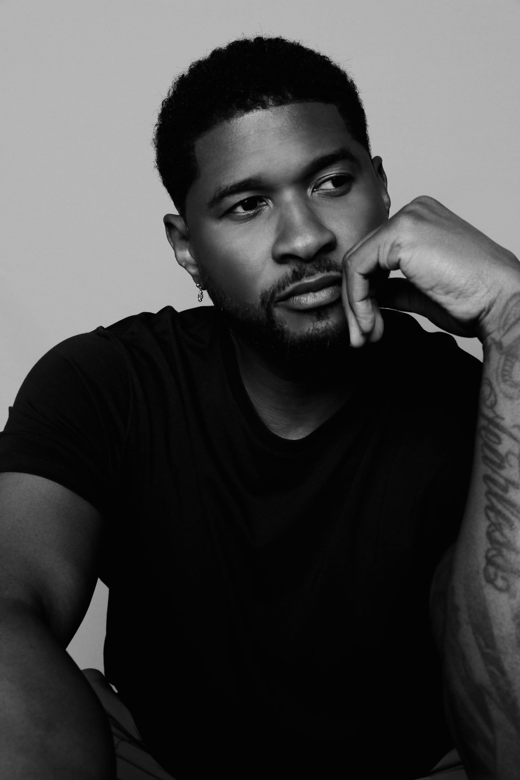 A black-and-white portrait of Usher, who is looking to the left with his right hand rested in a loose fist against his face. He is wearing a black T-shirt.