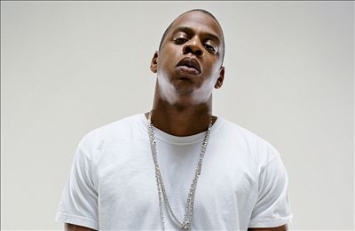 A picture of younger Jay-Z, wearing a white T-shirt and three silver chains. His head is tilted upwards with his eyes looking downward at the camera. The background is a white backdrop.