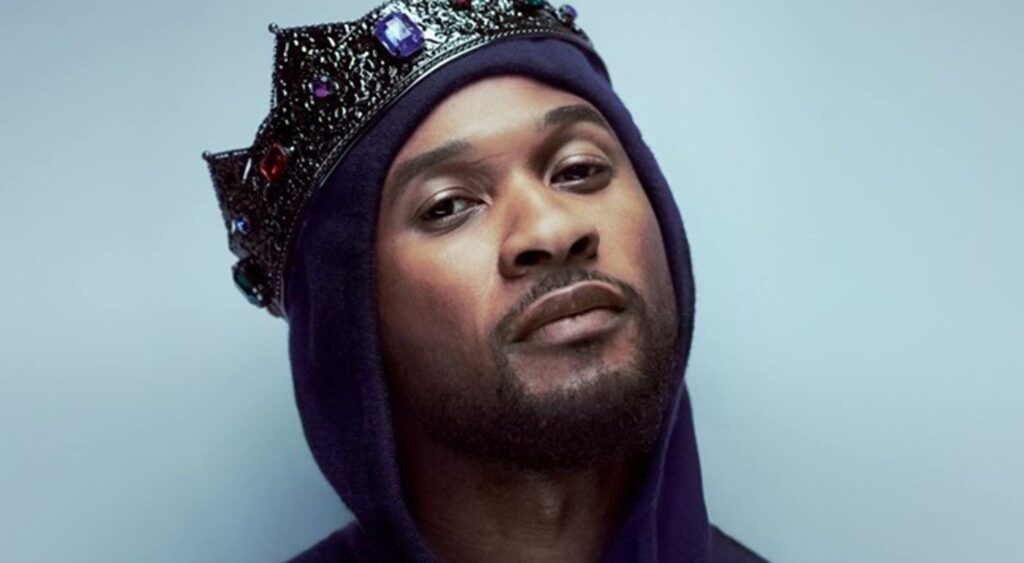 A portrait picture of Usher (only his face and neck), wearing a black crown and hoodie. His head is tilted upwards towards the right while looking straight at the camera in a suave manner. The backdrop is gray-blue.